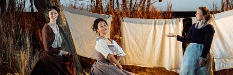 Three women are dressed in period costumes and standing in front of a field setting and sheets hanging up on a washing line. They are smiling and dancing.