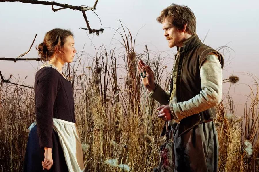 A man and a woman dressed in period costumes stand in a field setting, looking intensely at one another. The man is holding a medal in his hand.