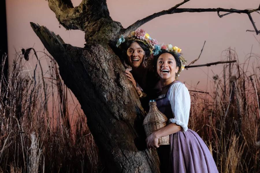 Two woman are dressed in period costumes with flower crowns on their head. One is holding a wicker jug and they are looking happy standing behind a tree.
