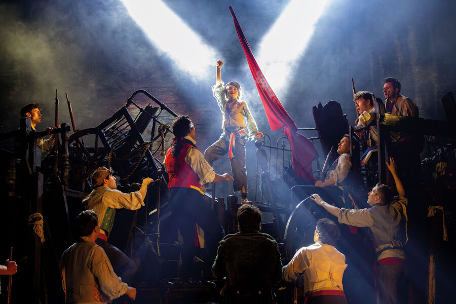 There are loads of people on a barricade with a young boy at the top with his fist raised. There is a red flag beside him and there's dramatic lighting which creates a tense atmosphere.