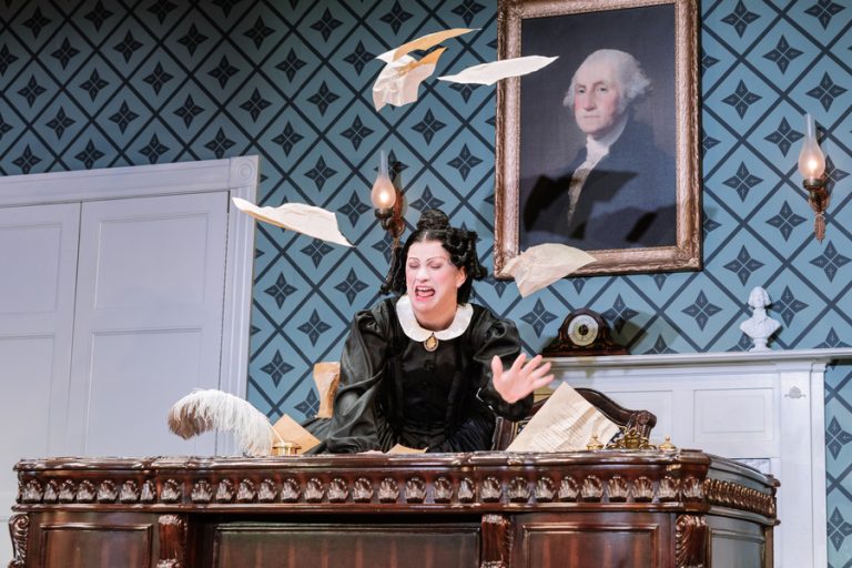 A production image of Jinkx Monsoon in Oh,Mary. She is throwing paper in the air on a desk and looks outraged.