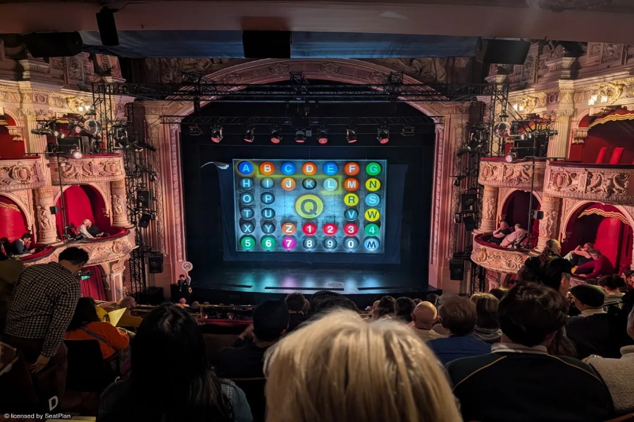 The stage at the Shaftesbury Theatre as seen from the Royal Circle. There is a brown proscenium arch above the stage and golden-brown boxes on either side of the stage. The safety curtain is down - it is designed to look like a New York subway map, with lots of colourful letters and numbers. In the centre is a large yellow Q. At the bottom of the image the backs of audience members heads can be seen. 