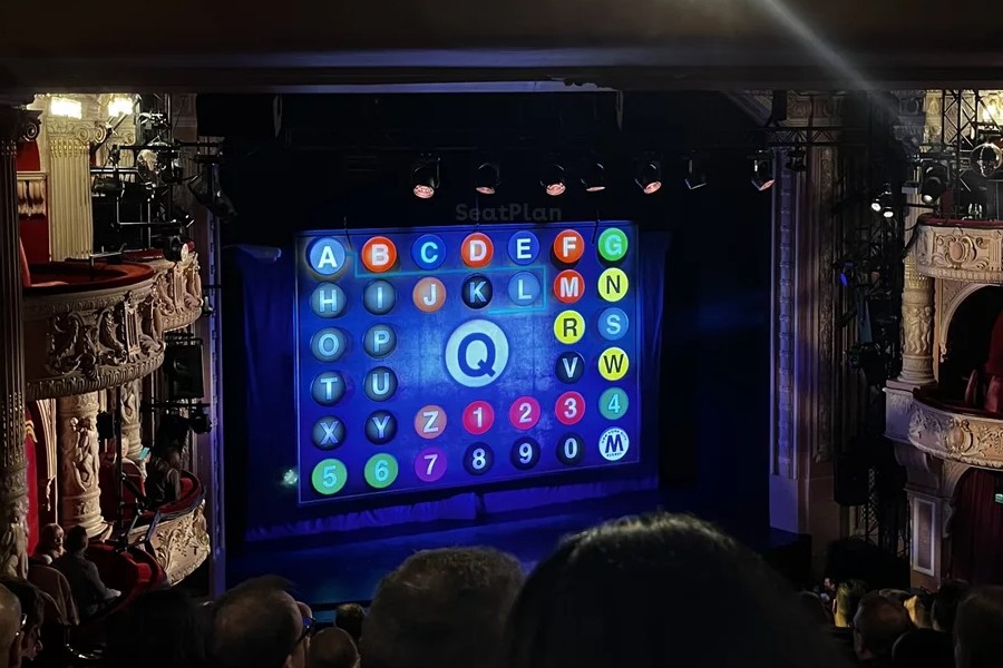 The stage at the Shaftesbury Theatre as seen from left of the Royal Circle. To the sides of the stage there are ornate boxes. The safety curtain is down - it is designed to look like a New York subway map, with lots of colourful letters and numbers. In the centre is a large yellow Q, which looks blue-ish in this image due to the lighting. At the bottom of the image the backs of audience members heads can be seen. 