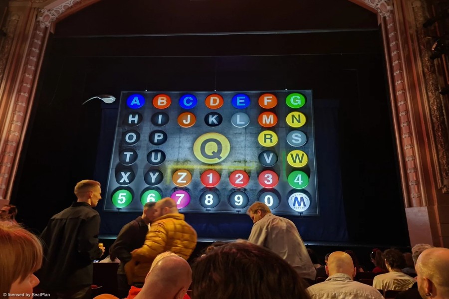 The stage at the Shaftesbury Theatre as seen from the Stalls. There is a brown proscenium arch above the stage. The safety curtain is down - it is designed to look like a New York subway map, with lots of colourful letters and numbers. In the centre is a large yellow Q. At the bottom of the image the backs of audience members heads can be seen. 