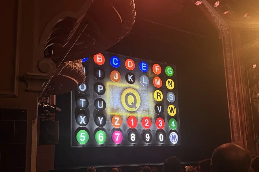 The stage at the Shaftesbury Theatre as seen from the left hand side of the Stalls. There is a brown proscenium arch above the stage, and the left side of the stage is slightly cut off at the very top by the bottom of one of the boxes above. The safety curtain is down - it is designed to look like a New York subway map, with lots of colourful letters and numbers. In the centre is a large yellow Q. At the bottom of the image the backs of audience members heads can be seen. 