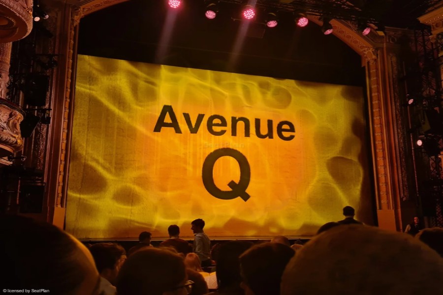 The stage at the Shaftesbury Theatre as seen from the Stalls . There is a brown arch above the stage. The safety curtain is down - it is bright yellow and fuzzy, with the words Avenue Q in black text in the centre. At the bottom of the image the backs of audience members heads can be seen. 