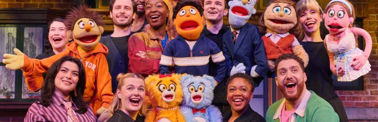 Avenue Q review header. Photo of the West End cast posing on stage with the puppets.