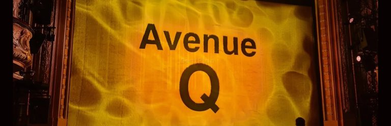 A seatview photo taken from the Shaftesbury Theatre stalls. A fuzzy yellow curtain with the words 'Avenue Q' in the centre in black.