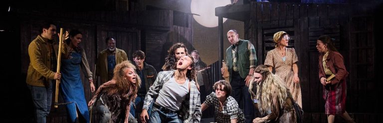 A group of performers are surrounding a man who is shouting out above him. There is a moon lit behind them.