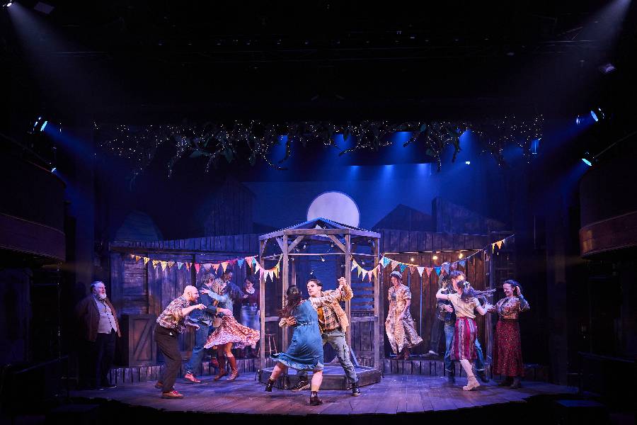 A group of performers are barn dancing in partners on the stage. Behind them is a stream of bunting and a lit up moon in the background. 