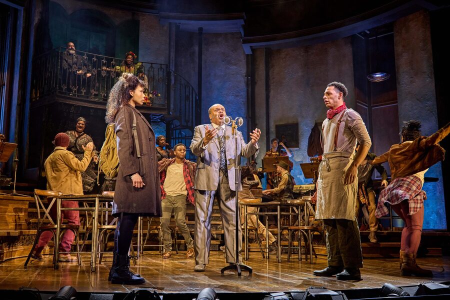 At the centre of this photo is a man in a grey suit singing into a vintage microphone. In front of him is a couple staring one another whilst an ensemble of performers dances around them.