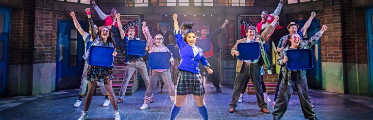 a group of performances are punching the air. the one in the centre is wearing a blue school uniform , whereas the performers around her are holding dinner trays.