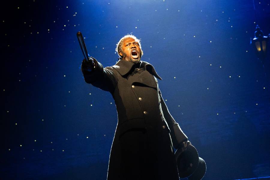 A performer in a dark coat is singing passionately, pointing a baton. The background is starry.