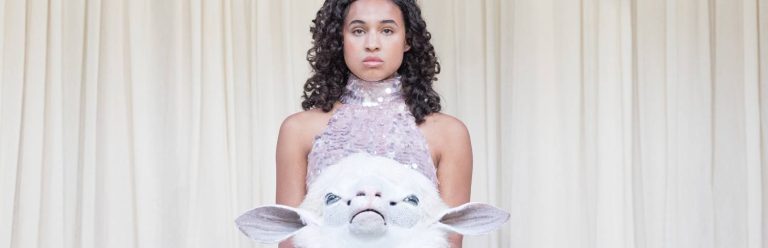 Girl with dark curly hair is stood holding a head of a lamb puppet. She is wearing a lilac sequinned dress and is stood in front of a white curtain.