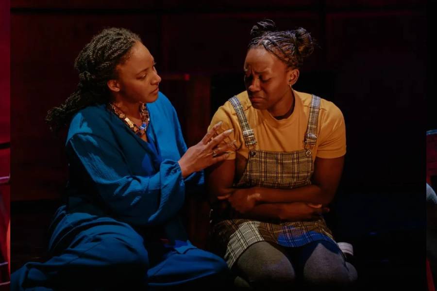 There are two women sat together. One on the left is wearing a blue outfit and is comforting the lady on the right, who looks upset. The woman on the right has her arms crossed and is a wearing a plaid dress and a yellow shirt.
