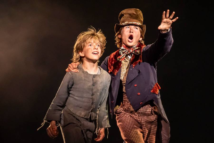 A performer with an old top hat and tails has his arms around another performer who is dressed in all grey.