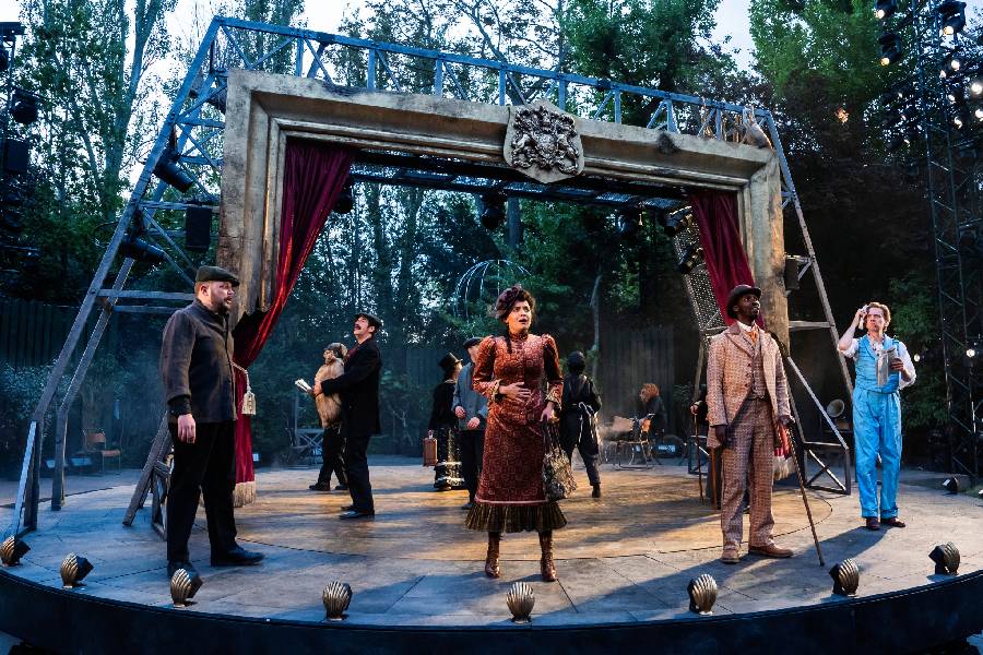 A group of actors are stood in period costumes in an outdoor theatre setting. Behind them is a large framed structure with red curtains. 
