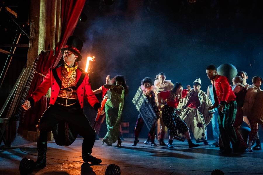 A circus ringmaster is standing on the left side of the photo. In the back is a group of performers holding various objects, including a torch. 