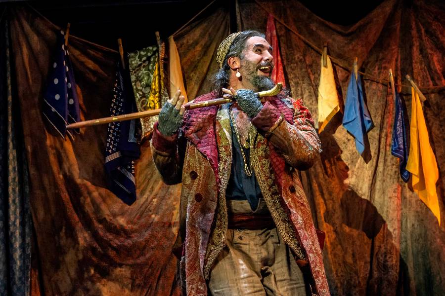 A theatre performer stands joyfully in a colourful costume with scarves hanging on a line behind him. He is holding a staff like a musical instrument in oliver.