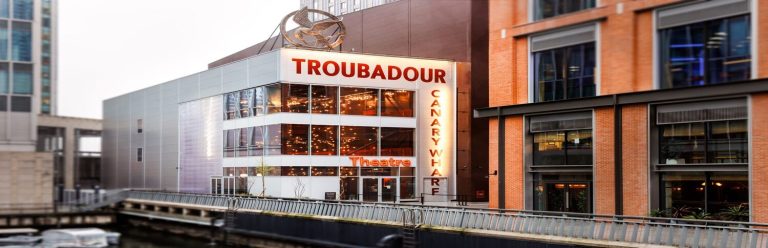 A modern theatre with "Troubadour Canary Wharf" sign on it. It has large doors and glass windows which reveal warm lights in it.